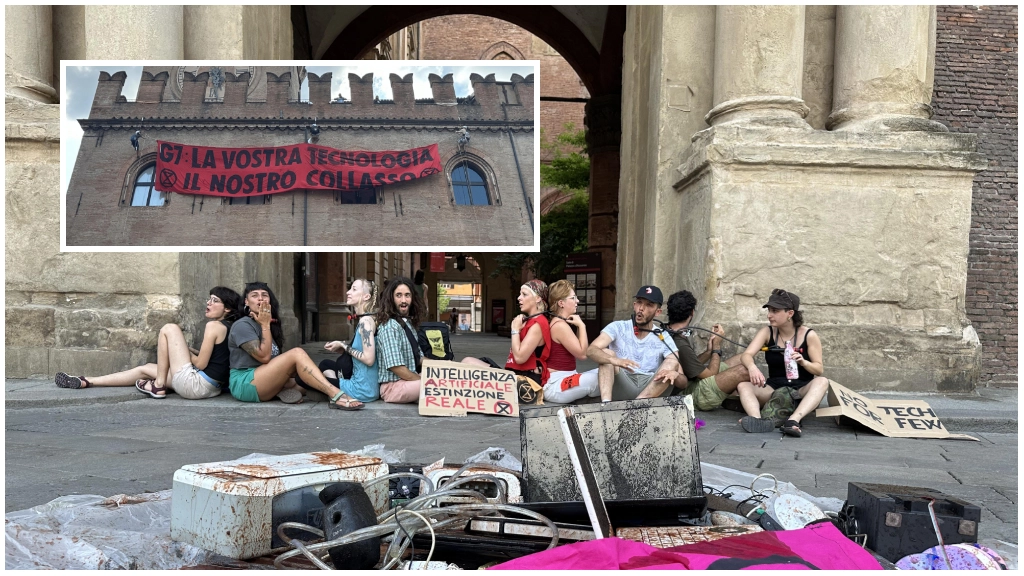 Gli attivisti che si sono incatenati davanti al Comune di Bologna; nel rettangolo lo striscione appeso sulla facciata di Palazzo d'Accursio